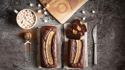 Two chocolate banana loafs and a cup of hot chocolate with marshmallow on a gray stone background