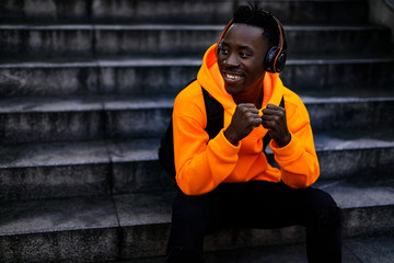 smiling african-american man in stylish orange hoodie sweatshirt in wireless headphones listening music and sitting on stairs. copy space