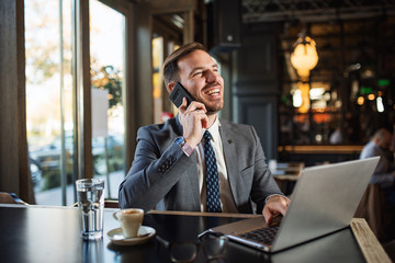 Handsome smiling businessman using phone and laptop in cafe bar