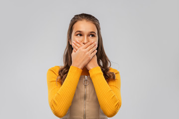 emotion, expression and people concept - scared teenage girl closing her mouth by hands over grey...