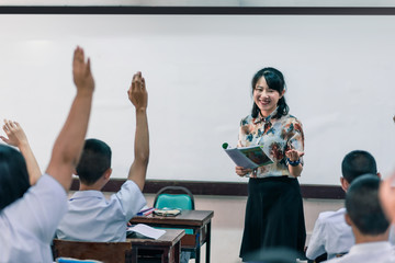 A smiling Asian female high school teacher teaches the white uniform students in the classroom by asking questions and then the students raise their hands for answers.
