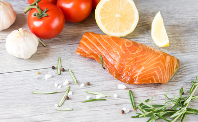 Grilled salmon steak on vintage wooden board with fork, olive oil, vegetables, herbs and spices on dark concrete background. Raw salmon fillet with vegetables, spices and lemon on wooden table.