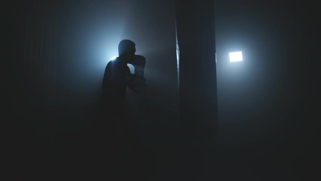 Alone boxer hits punching bag in dark gym in slow motion. Young man training indoors. Strong athlete boxing in smoky studio. Sport concept. Low key. Medium shot in 4K, UHD