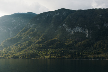 A view of the hill at Lake Bohinj. The green and wild mountains that surround the lake belong to Triglav National Park, Slovenia. Sunrise on the lake. Calm water in the foreground.