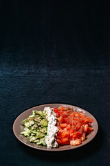 Simple salad of cucumbers and tomatoes with sour cream flavored with salt and black pepper on a dark background