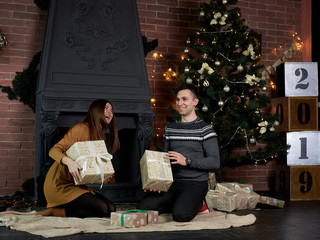 Handsome brunette man in grey sweater and young woman in mustard yellow knitted dress holding presents. Couple in love sitting on carpet by fireplace and Christmas tree in dark room with brick wall.