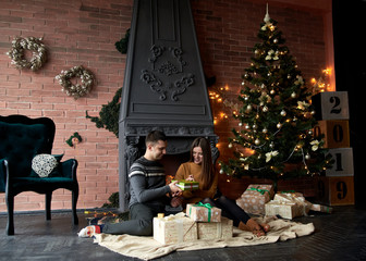 Handsome brunette man, wearing grey sweater giving present to pretty woman in mustard yellow knitted dress. Young couple in love sitting by fireplace near Christmas tree in dark room with brick wall.