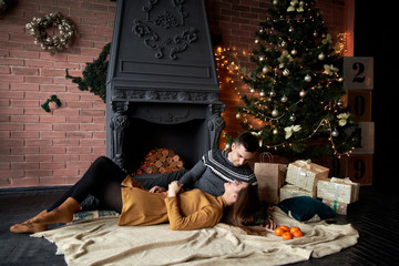 Handsome brunette man in grey sweater and young woman in mustard yellow knitted dress lying on the floor carpet near Christmas tree, embracing. Couple in love by fireplace in dark room with brick wall
