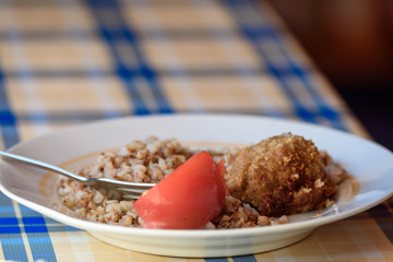 Plate of crumbly buckwheat with steam patty and tomato on the table.