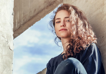 Lovely girl in dark clothes sits on concrete block in industrial zone. Face close-up