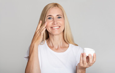 Skin care products. Happy lady applying face cream on cheek