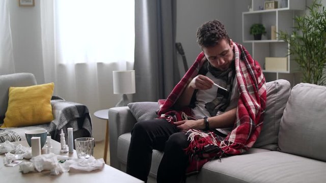 A sick man is measuring the temperature at home on the sofa