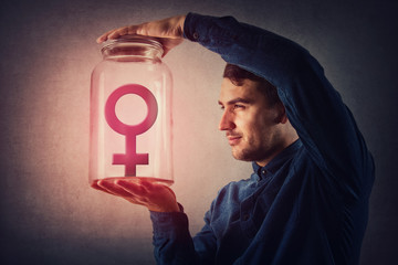 Young male, malefic look holding a glass jar with female gender symbol inside as captive. Man pretending to be superior to woman, gender gap idea, sex inequality concept. Social issue, discrimination.