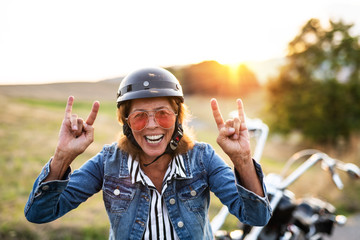 A cheerful senior woman traveller with motorbike in countryside at sunset.