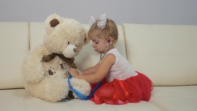 Little girl plays a doctor with a stethoscope and a teddy bear. girl sitting on the couch. The concept of children's fantasies and games. Training as a doctor