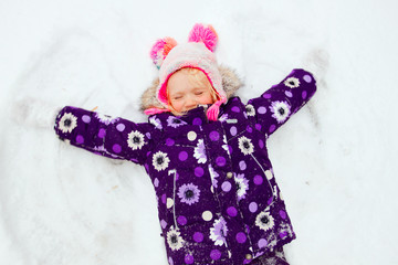 Cute toddler girl wearing bright color snow jacket and funny hat making snow angel outdoors in a cold winter day 