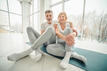 Sporty senior couple sitting on the mat in the white fitness gym looking at camera and smiling. Sports and healthy lifestyle.