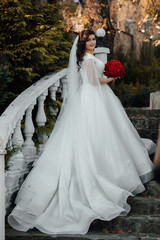 Naklejka na ściany i meble Beautiful sensual young bride in long white wedding dress and veil the bride on the stairs and holding bouquet outdoor. Charming autumn bride. Beautiful woman with professional make up and hair style.