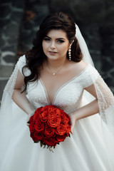 Charming autumn bride. Beautiful woman with professional make up and hair style. The bride holds a bouquet of red roses.  Fashion portrait of gorgeous woman