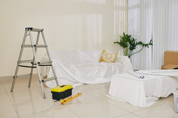 Furniture covered with white fabric, ladder, toolbox and construction plans in room under renovation