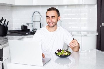 Fototapeta na wymiar Optimistic man wearing casual clothing eating breakfast at home while using laptop