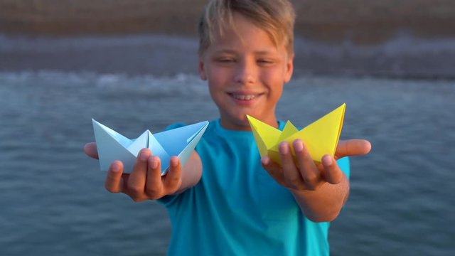 Closeup view of cute handsome white kid holding two paper bright blue and yellow colors ships standing at sunny morning beach. Focus slides from toys to face and backward. Real time 4k video footage.
