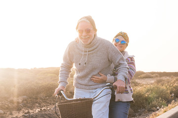 Playful and youthful senior caucasian peope couple enjoy a ride with bike together laughing and smiling -    concept of happiness and joyful intoegther forever life with love