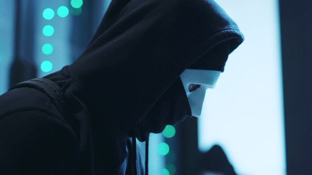 Stressful hacker in mask breaking server in data center looking around nervously. Close-up computer spy coding a malware program to steal database information.