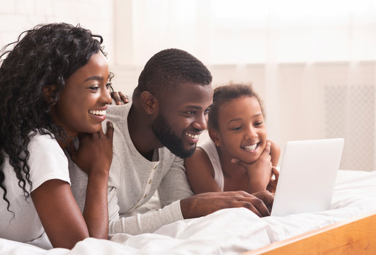 Joyful black family of three using laptop in bed together