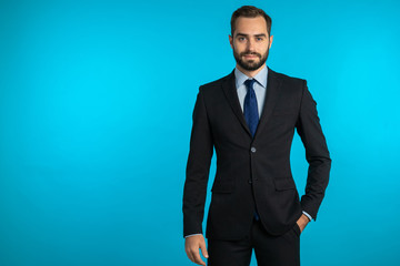 Copy space. Portrait of young successful confident businessman with beard isolated on blue studio background. Man in business suit looking to camera and smiling. Portraiture of handsome guy. - Powered by Adobe