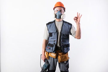 Builder with a drill puncher, in a helmet, and a respirator. Shows okay sign