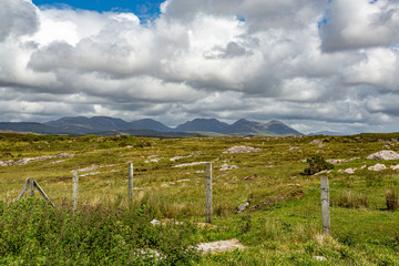 Fences in the Irish countryside with green grass and limestone rock mountains in the background at Derrigimlagh Bog, spring day with cloudy sky in County Galway, Ireland