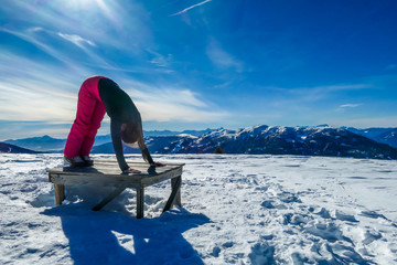 A girl in skiing outfit standing in a downward facing dog's pose on a bench, surrounded by powder snow in Bad Kleinkirchheim, Austria. Girl is practising yoga in the nature, finding inner peace.