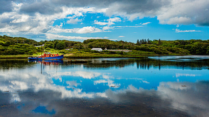 Clifden bay with clouds reflecting in the water and a boat anchored in the water at high tide, surrounded by green vegetation, spring day with a blue sky and white clouds Ireland