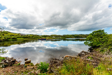 Clifden Bay seen from the shore with clouds reflecting in the water, a ship anchored at high tide, surrounded by green vegetation, spring day with a blue sky and white clouds in Clifden, Ireland