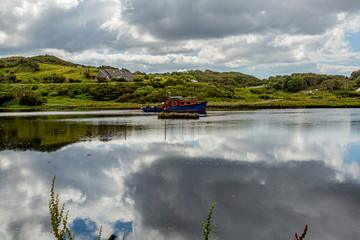 Boat anchored in the middle of Clifden bay at high tide, clouds reflected in the water, surrounded by green vegetation, spring day with a blue sky and white clouds in Clifden, Ireland