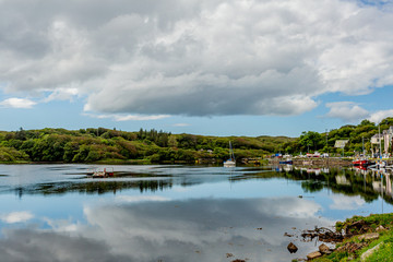 Calm waters, reflection and boats anchored on the shore at Clifden Harbor at high tide, sunny spring day with a blue sky and white clouds in Clifden, Ireland