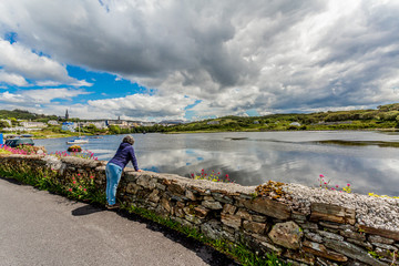 Mature woman leaning on a stone fence looking at the pier in the port of Clifden with anchored...