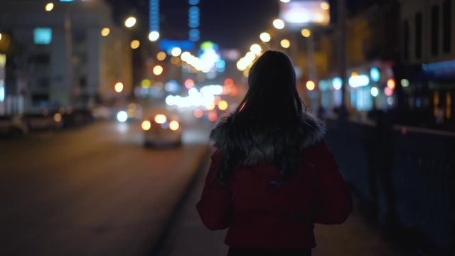 Young woman with hair in the tail and red jacket with fur walking along road in city street past the lights of passing cars. Against background of the night city bokeh, back view of dark silhouette.