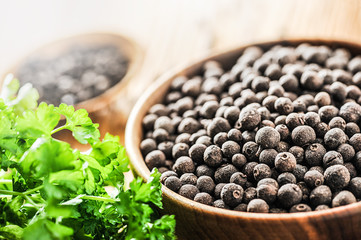 Black dried pepper in wooden bowl with green leaves background. Various peppers on wooden table.