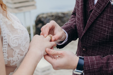 the bride and groom put on each other rings, a burgundy suit and a beige wedding dress, cloudy day, a ceremony by the sea, hands close-up