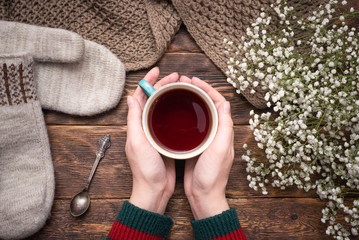 Female hands in sweater with a cup of hot tea above wooden table background.