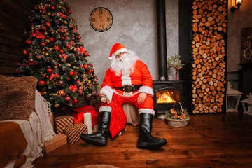 Thoughtful Santa Claus sits in a chair inside of his house with bag full of gifts and get ready for christmas greetings. Beautiful home decoration