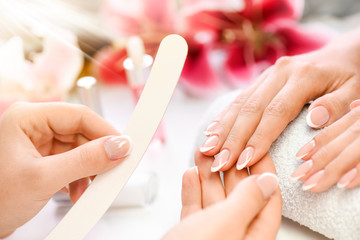 Woman beautician using  a nail file. Professional and beautiful hands with nails care manicure applying in luxury salon. Pink red flowers background.