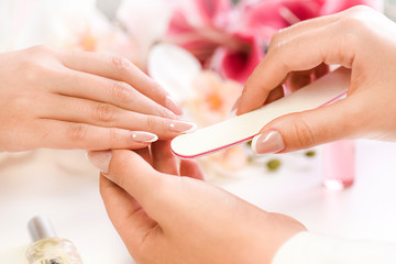 Obraz na płótnie Canvas Woman beautician using a nail file. Professional and beautiful hands with nails care manicure applying in luxury salon. Pink red flowers background.