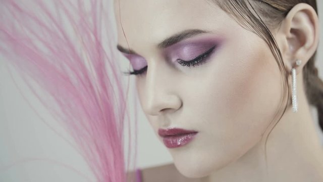 Glamorous young girl with pink make-up on her face with pink lip gloss. Luxurious pink feathers near the girl's face. Advertising perfume. Young woman hiding face with a pink feather.