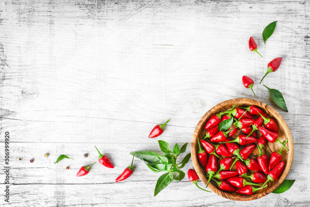 Wall mural Red fresh chili peppers in wooden bowl on white old table. Spicy or hot small pepper with green leaves top view and copy space.