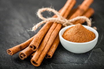 Cinnamons dried sticks on dark stone table. Cinnamon ginger powder in white bowl on black board.