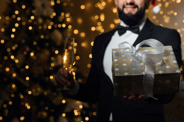 Photo of one handsome new year man with beard in red santa claus christmas hat in studio. The guy holds a glass of champagne and a gift. Merry Christmas and happy New Year! 