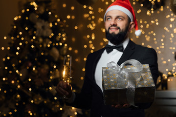 Photo of one handsome new year man with beard in red santa claus christmas hat in studio. The guy holds a glass of champagne and a gift. Merry Christmas and happy New Year! 